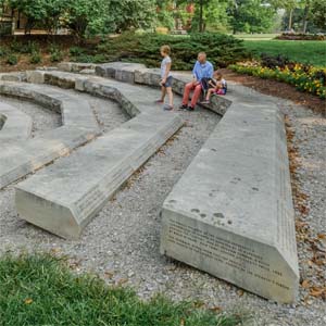 A man and a young girl sit on a stone bench, which is part of the Freedom Summer Memorial on Western Campus of Miami University. A second young girl stands on an adjacent stone bench, and all three people look toward the center of the memorial.